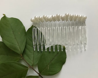May include: A clear plastic hair comb with white pearl beads on the top. The comb is lying on a white surface with a green leaf branch next to it.