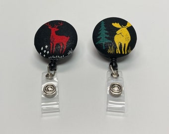 Può includere: Due porta badge retrattili neri con un designo di cervo rosso e alce giallo. Il cervo è in piedi in un bosco con alberi bianchi e verdi. L'alce è in piedi in un bosco con alberi verdi e gialli.