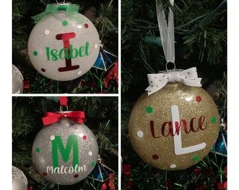 May include: Three personalized Christmas ornaments with glitter and colorful dots. The ornaments are silver, gold, and white with the names "Isabel", "Lance", and "Malcolm" written on them.
