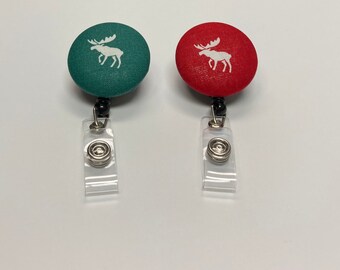 Può includere: Due porta badge retrattili con un design a forma di alce. Un porta badge ha un bottone in tessuto verde con un alce bianco, l'altro ha un bottone in tessuto rosso con un alce bianco.