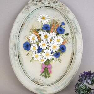 May include: A white oval frame with a distressed finish, featuring a 3D floral arrangement of white daisies and blue cornflowers with wheat stalks, tied with a pink ribbon.