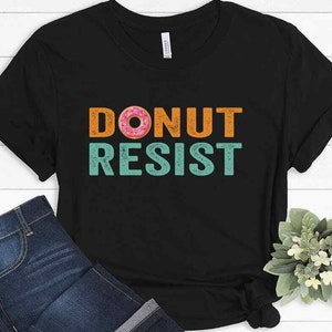 Pode incluir: Camiseta preta com um gr&aacute;fico de estilo vintage que diz "DONUT RESIST" com um donut rosa e branco no meio da palavra "DONUT".