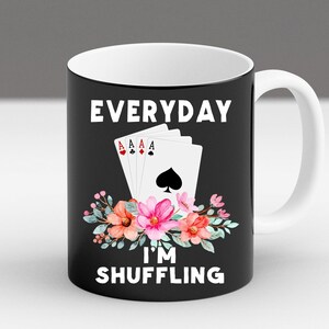 Pode incluir: Caneca de cerâmica preta com alça e interior brancos. A caneca apresenta o texto "EVERYDAY I'M SHUFFLING" em branco, com cartas de baralho e detalhes florais. O design inclui quatro ases e flores coloridas.