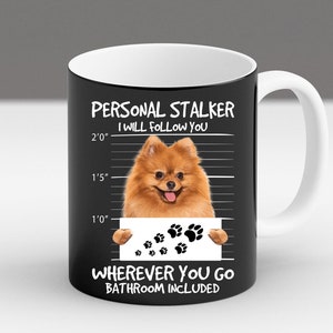 May include: Black ceramic mug with a white interior and handle. The mug features a graphic of a Pomeranian dog with the text "PERSONAL STALKER I WILL FOLLOW YOU WHEREVER YOU GO BATHROOM INCLUDED".