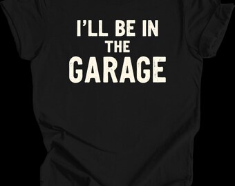 May include: Black t-shirt with the text "I'LL BE IN THE GARAGE" in a distressed, off-white font. The shirt is made of a soft material and has short sleeves. The text is centered on the front of the shirt.