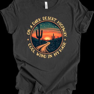 Puede incluir: Camiseta gris oscuro con un dise&ntilde;o circular que representa una escena de carretera des&eacute;rtica. El dise&ntilde;o incluye una carretera que conduce a una puesta de sol, cactus y el texto "ON A DARK DESERT HIGHWAY COOL WIND IN MY HAIR."