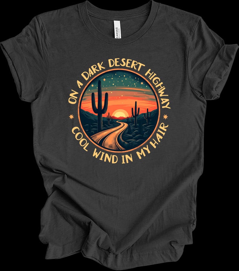 Puede incluir: Camiseta gris oscuro con un dise&ntilde;o circular que representa una escena de carretera des&eacute;rtica. El dise&ntilde;o incluye una carretera que conduce a una puesta de sol, cactus y el texto "ON A DARK DESERT HIGHWAY COOL WIND IN MY HAIR."