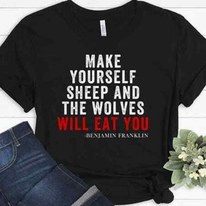 May include: Black t-shirt with the quote "MAKE YOURSELF SHEEP AND THE WOLVES WILL EAT YOU" in white and red text. The quote is attributed to Benjamin Franklin. The shirt is paired with blue jeans.
