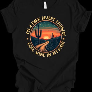 Puede incluir: Camiseta negra con un dise&ntilde;o circular que representa una escena de carretera des&eacute;rtica. El dise&ntilde;o incluye una carretera que conduce a una puesta de sol, cactus y el texto "ON A DARK DESERT HIGHWAY COOL WIND IN MY HAIR."