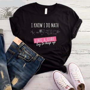 May include: Black t-shirt with the text "I know I do math like a girl try to keep up" in white. The text is accompanied by a pink rectangle with the words "like a girl" in white. The shirt also features various mathematical equations and diagrams in white.