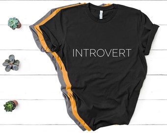 May include: Black t-shirt with the word "INTROVERT" printed in white on the front. The shirt is folded and layered with a mustard yellow t-shirt and a gray t-shirt.