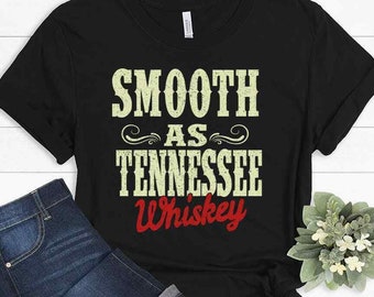 Può includere: T-shirt nero con la scritta "Smooth as Tennessee Whiskey" in crema e rosso, in stile vintage. Il testo è impreziosito da decorazioni. La maglietta è abbinata a un paio di jeans blu.