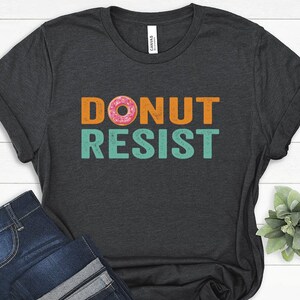 Pode incluir: Uma camiseta cinza escura com o texto "DONUT RESIST" em laranja e turquesa. Um donut rosa e laranja est&aacute; no meio da palavra "DONUT".