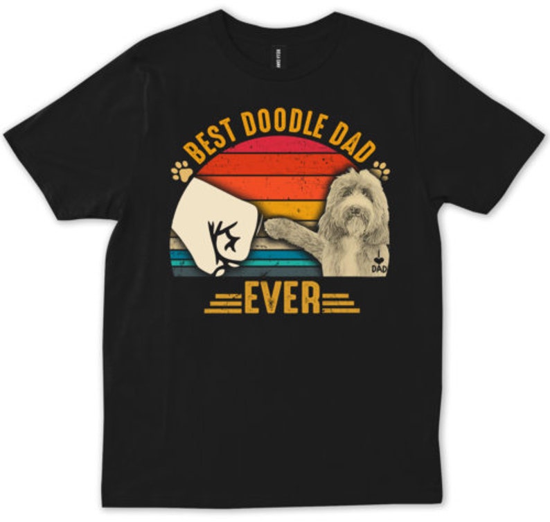 May include: Black t-shirt with a graphic that says "Best Doodle Dad Ever." The design features a fist bump with a dog's paw and a doodle dog silhouette against a sunset background. The word "DAD" is on the dog's chest.