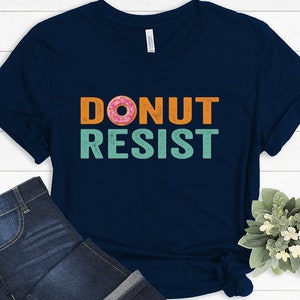 Pode incluir: Uma camiseta azul marinho com o texto "DONUT RESIST" em laranja e turquesa com um gr&aacute;fico de donut rosa e laranja.