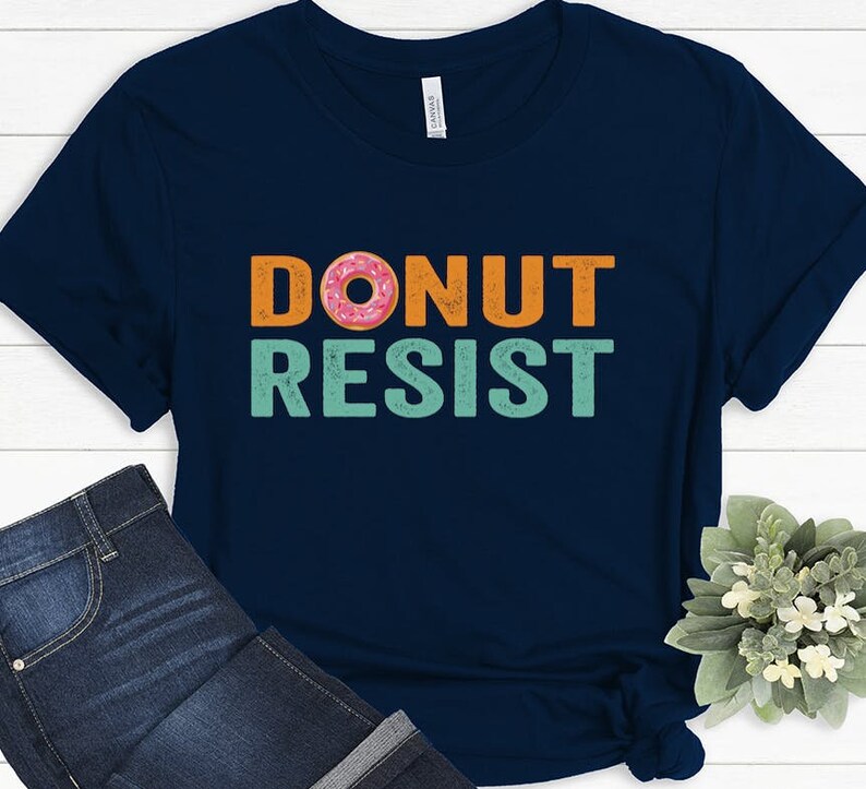 Pode incluir: Uma camiseta azul marinho com o texto "DONUT RESIST" em laranja e turquesa com um gr&aacute;fico de donut rosa e laranja.