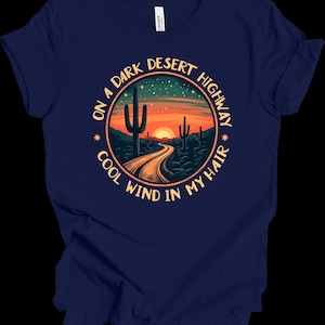 Puede incluir: Camiseta azul marino con un dise&ntilde;o de paisaje des&eacute;rtico. El dise&ntilde;o incluye una carretera que conduce a una puesta de sol, cactus y el texto "ON A DARK DESERT HIGHWAY COOL WIND IN MY HAIR" en un patr&oacute;n circular. La camiseta es de material suave.