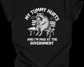 May include: Black t-shirt with a white graphic of a raccoon sitting on a bench. The text "My tummy hurts and I'm mad at the government" is printed on the shirt.
