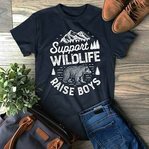 Könnte beinhalten: Ein dunkelblaues T-Shirt mit einem weißen Grafikdesign, das einen Bären, Berge und Bäume zeigt. Der Text auf dem Shirt lautet "Support Wildlife Raise Boys".