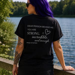 Könnte beinhalten: Ein schwarzes kurzärmeliges T-Shirt mit weißem Text auf der Rückseite, der lautet: DEAR PERSON BEHIND ME: YOU ARE STRONG. incredible. and enough. Always remember. Das Design enthält eine kleine weiße Herzumrandung neben dem Wort STRONG.