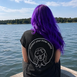 Könnte beinhalten: Ein schwarzes kurzärmeliges T-Shirt mit einer weißen kreisförmigen Grafik auf der Rückseite. Das Design zeigt eine Strichzeichnung eines Gesichts mit einer Schlafmaske, die mit Sternen, einem Mond und einem Herz verziert ist, sowie den Text "Protect your inner world."