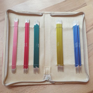 Könnte beinhalten: Eine beige Reißverschlusstasche mit sechs Reihen Stricknadeln. Die Nadeln sind in vier Farben: rosa, rot, grün, gold und blau.