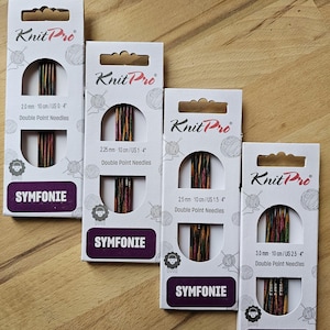 Könnte beinhalten: Vier Packungen Knit Pro Symfonie Doppelspitznadeln in verschiedenen Größen. Die Nadeln sind aus Holz und haben eine farbenfrohe, marmorierte Oberfläche. Die Packungen sind weiß mit schwarzem Text und einem Schaf-Logo.