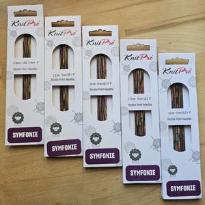 Könnte beinhalten: Fünf Packungen Knit Pro Symfonie Doppelspitzennadeln in verschiedenen Größen. Die Packungen sind weiß mit einem schwarz-weißen Schaflogo und dem Text "Knit Pro" und "Symfonie". Die Nadeln sind aus Metall und haben eine farbenfrohe, marmorierte Oberfläche.