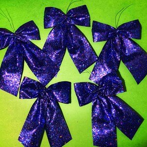 May include: Five purple glitter bows with a green background. The bows are made of fabric and have a shiny, sparkly finish.
