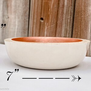 May include: A white concrete bowl with a 7 inch diameter and 2 inch depth. The bowl has a light orange rim.