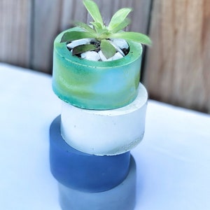 May include: A stack of three concrete planters in shades of blue and green. The top planter has a small succulent plant growing in it.