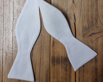 May include: White bow tie made from a textured fabric. The bow tie is folded and laid flat on a wooden surface.