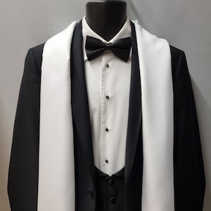 May include: A black tuxedo jacket with a white satin shawl collar and a white bow tie. The jacket is worn over a white dress shirt and a black waistcoat. A white fringed scarf is draped around the neck.