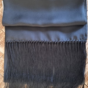 May include: A black scarf with a silky texture and a fringe trim. The scarf is folded and lies on a wooden surface. The fringe is long and hangs down from the bottom edge of the scarf.