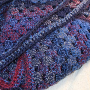 May include: A close-up of a crocheted scarf in shades of blue and burgundy. The scarf is made with a textured stitch and has a soft, fuzzy appearance.