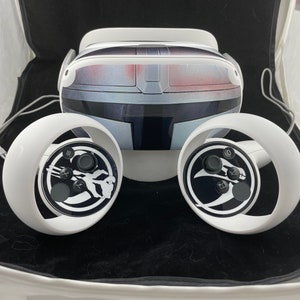 May include: A white virtual reality headset with two white controllers. The headset has a black and gray design with a textured surface. The controllers have a black and white design with a circular button and a joystick.