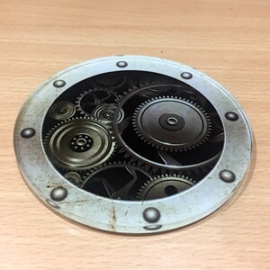 May include: A round, white coaster with a circular window showing a design of interlocking gears in shades of silver and gold against a black background.