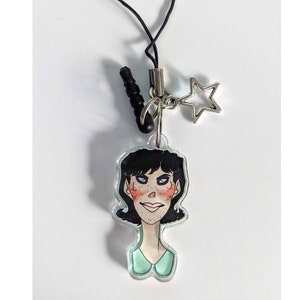 May include: A cell phone charm featuring a cartoon illustration of a woman with black hair and rosy cheeks. The charm has a silver-colored border and a small star-shaped charm. A black cord is attached to the top.