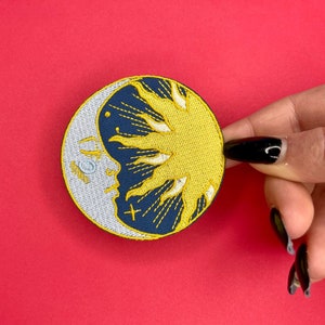 May include: An embroidered patch featuring a yellow sun and white crescent moon, with a navy blue background. The sun and moon overlap and have a stylised design.