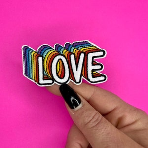 Op de afbeelding: Een regenboogkleurige strijkpatch met het woord "LOVE" in witte letters. De patch is in 3D-stijl, met de letters die op elkaar lijken te liggen.