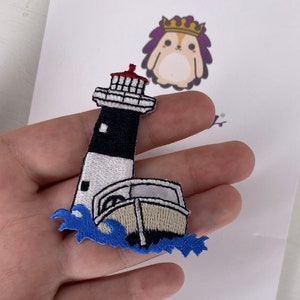 Könnte beinhalten: Ein gestickter Bügel-Patch mit einem weißen Leuchtturm mit schwarz-rotem gestreiftem Oberteil, einem kleinen weißen Boot und blauen Wellen.