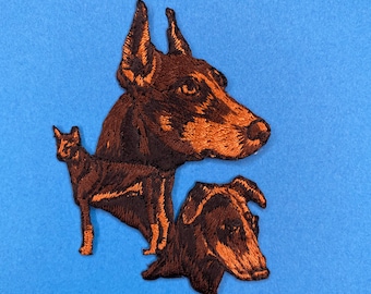 May include: Embroidered patch featuring three brown dogs. The top dog is a Doberman Pinscher with its head turned to the side. The other two dogs are smaller and are partially obscured by the Doberman.