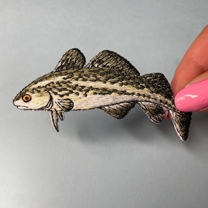 Peut inclure: Patch thermocollant brodé représentant un poisson brun et blanc avec des détails noirs. Le poisson a un gros œil et une bouche légèrement ouverte.