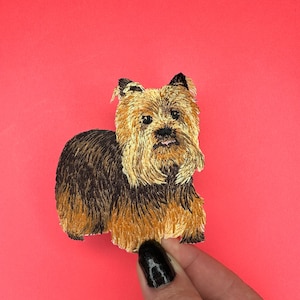 May include: Embroidered patch of a brown and black Yorkshire Terrier dog with a light brown face. The dog is looking directly at the camera.