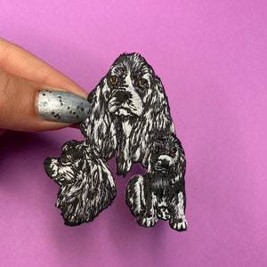 May include: Embroidered patch featuring three black and white cocker spaniels. The largest spaniel is in the center with its head facing forward. The other two spaniels are on either side of the largest spaniel, with their heads turned to the side.