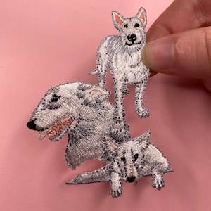 May include: Three white embroidered iron-on patches of dogs. The patches are in different poses. The first patch is of a dog standing with its head tilted. The second patch is of a dog lying down with its head turned to the side. The third patch is of a dog sitting with its head tilted.