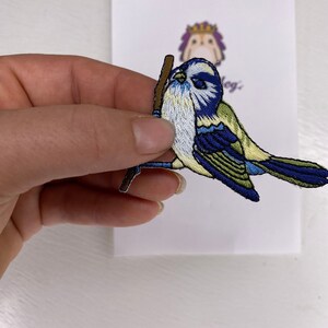 Könnte beinhalten: Ein gestickter Bügelaufkleber mit einem blauen Vogel, der einen Stock in seinem Schnabel hält. Der Vogel ist blau, weiß und grün mit einem braunen Stock.