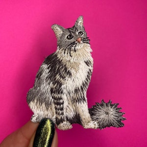 Könnte beinhalten: Gestickter Bügel-Patch mit einem grau-weißen Kätzchen mit einem flauschigen Schwanz. Die Katze sitzt und schaut nach rechts.