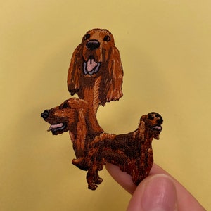 Puede incluir: Un parche bordado marrón y negro de un perro con la lengua fuera. El perro está sentado con la cabeza inclinada hacia un lado. El parche está hecho de fieltro y tiene un borde cosido.