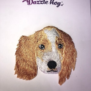 May include: Embroidered patch of a brown and white dog's face. The patch is made of fabric and has a detailed design. The dog has brown fur, a white muzzle, and black eyes.
